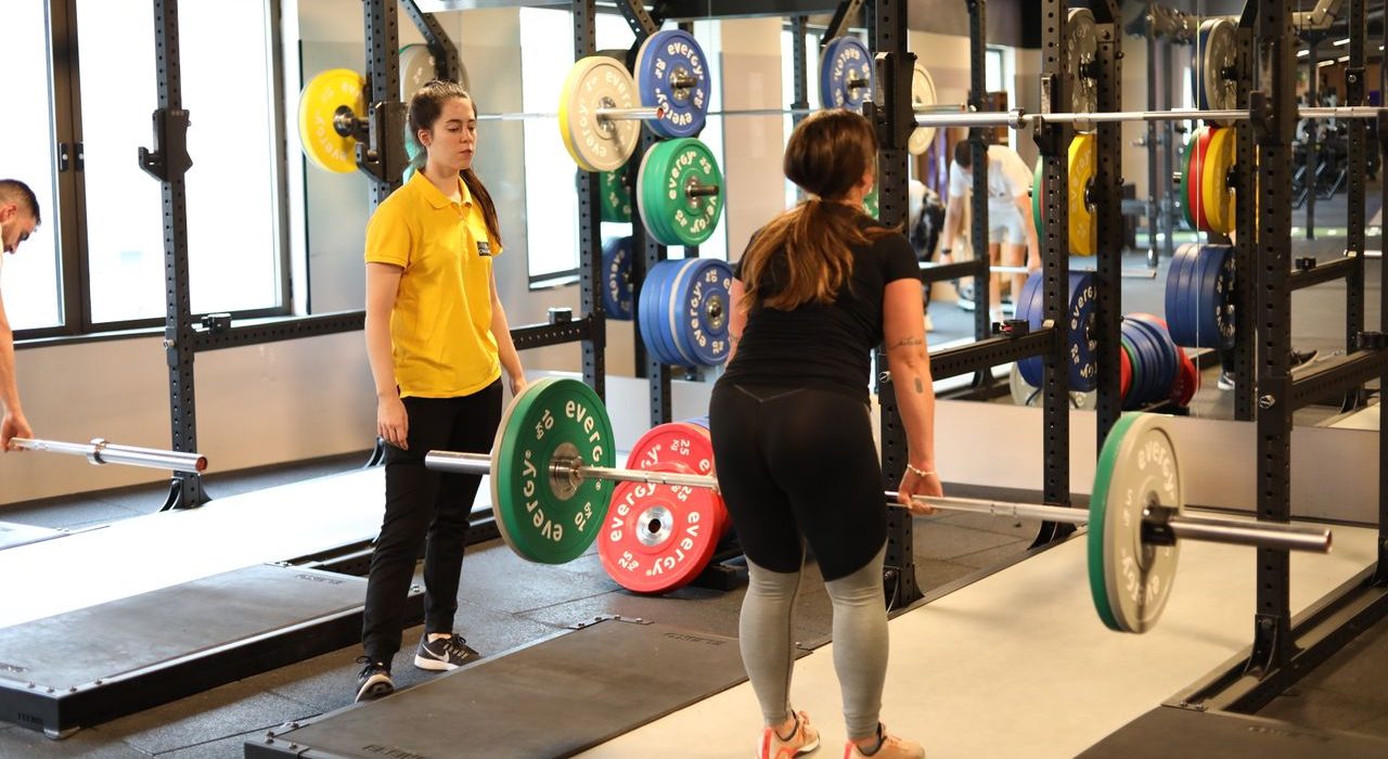 Two women weightlifting in a modern gym with colorful plates, showcasing 8-weeks get ripped fitness training.