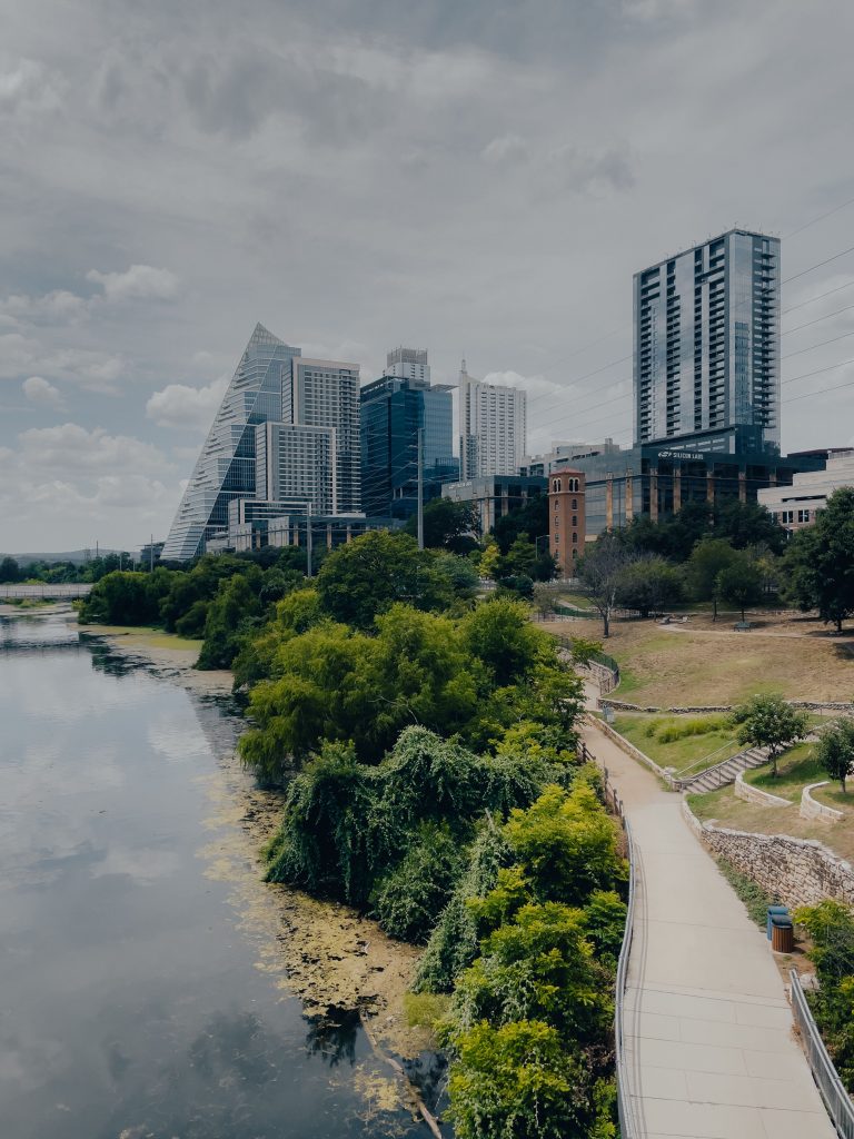 A serene urban cityscape featuring skyscrapers by a riverside pathway under a cloudy sky.